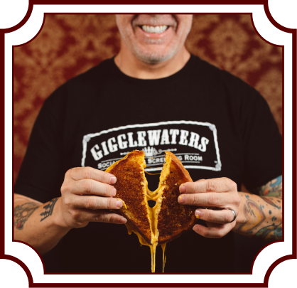 Smiling person wearing a Gigglewaters t-shirt holding a grilled cheese sandwich with melted cheese stretching down, against a decorative burgundy wallpaper background. Image has a vintage-style decorative frame border.
