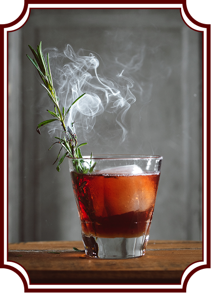 A red-amber cocktail in a rocks glass on a wooden surface, garnished with fresh rosemary that's smoking, creating an artistic plume of white smoke rising above the drink. The image has a vintage-style decorative frame border.